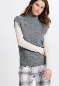 Maglione senza maniche grigio in maglia con colletto alto, caratterizzato da una finitura testurizzata. Indossato sopra una camicia a maniche lunghe chiara e pantaloni a quadri.