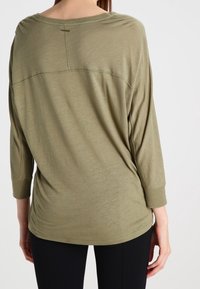 Olivově zelený, dlouhý rukáv, volně střižený top s měkkou texturou a kulatým výstřihem. Obsahuje viditelné švy na zadní straně.