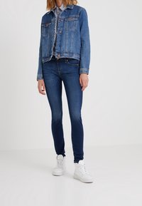 Chaqueta de mezclilla en azul medio con botones de cobre, dos bolsillos en el pecho y un diseño corto. Combinada con jeans ajustados azul oscuro y zapatillas blancas.