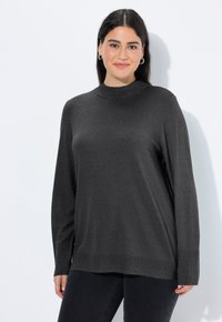 Frau mit dunkelhaarigen, die silberne Creolen trägt und einen dunkelgrauen Langarmpullover anhat, steht vor einem weißen Hintergrund.