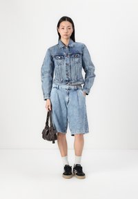 Chaqueta de denim y pantalones cortos en un lavado azul claro. La chaqueta tiene un cierre de botones y bolsillos en el pecho. Se combina con zapatillas negras y un bolso marrón.
