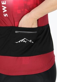 Haut de sport rouge et noir avec un corps texturé, doté d'une poche zippée et d'un logo de montagne blanc sur la section noire.
