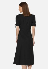 Schwarzes Midikleid mit Puffärmeln, ausgestattet mit Bindebändern an den Ärmeln. Ein tailliertes Oberteil, das in einen lockeren Rock übergeht, aus leichtem Stoff gefertigt.