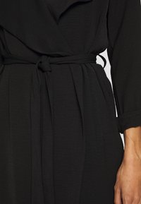 Czarna sukienka kopertowa z fakturowanej tkaniny, z paskiem wiązanym w talii oraz długimi rękawami z podwiniętymi mankietami.