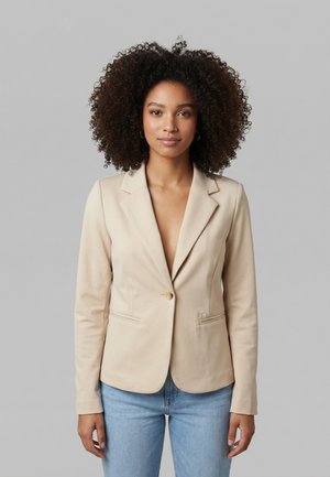 Mujer con cabello rizado que lleva un blazer beige de un solo botón y jeans azul claro, de pie contra un fondo gris.