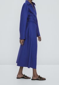 Långärmad, djupt blå skjortklänning med bälte i midjan, krage och A-linjeformad siluett, matchad med svarta platta sandaler.