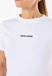 Biela bavlnená tričko s okrúhlym výstrihom a krátkymi rukávmi. S čiernym logom "PEGADOR" potlačeným na hrudi. Mäkká textúra, jednoduchý dizajn.