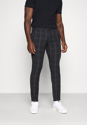 Man draagt een zwart shirt met korte mouwen, slank zittende donkergrijze geruite broek en witte Adidas sneakers, staand tegen een effen achtergrond.