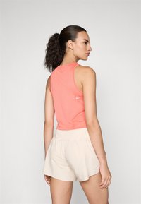 Koralkasta atletska majica bez rukava s teksturiranim mrežastim dizajnom i Nike logom, u kombinaciji sa svijetlim bež šortsevima, prikazana sa stražnje strane.