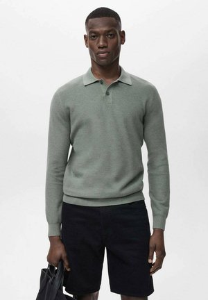 Polo tricot à manches longues, de couleur vert clair, avec un col, deux boutons et des bords côtelés. Porté avec un short noir et un sac noir. Fond uni.