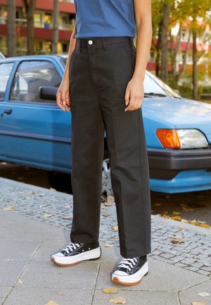 Schwarze, weit geschnittene Hosen aus robustem Stoff, mit Knopfverschluss und Seitentaschen. Kombiniert mit schwarz-weißen Turnschuhen.