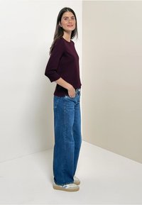 Bordowy longsleeve w połączeniu z niebieskimi spodniami o szerokich nogawkach, uzupełniony beżowymi i białymi trampkami, na jasnym tle.