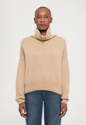 Beige knit sweater with a high collar, half-zip closure, and ribbed cuffs. Features a textured pattern and a relaxed fit. Worn with blue jeans.