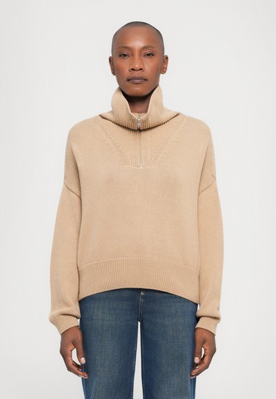 Maglione lavorato a maglia beige con colletto alto, chiusura a mezza zip e polsini a coste. Presenta un motivo testurizzato e una vestibilità rilassata. Indossato con jeans blu.