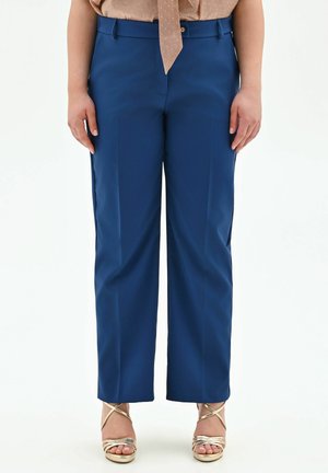 Donna che indossa pantaloni blu sartoriali, camicetta beige con dettaglio a fiocco e sandali dorati con cinturini e tacco alto, in piedi su sfondo bianco.