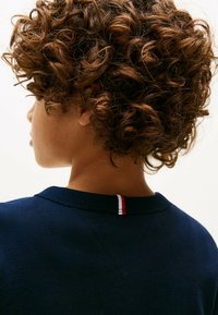 Felpa blu navy con collo tondo, caratterizzata da un accento a strisce rosso, bianco e blu al colletto; materiale in cotone morbido e testurizzato; capelli ricci visibili.