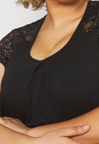 Top negro de manga corta con detalles de encaje en los hombros, cuello de pico y pliegue frontal, fabricado en material suave y elástico.