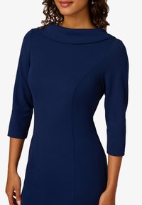 Granatowa sukienka z wysokim kołnierzem i rękawami do trzech ćwiartek. Wykonana z gładkiego materiału, z dopasowanymi szwami, które podkreślają sylwetkę.
