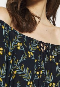 Blusa oscura de flores con escote asimétrico, con patrones de flores amarillas y rosas, tela texturizada y detalle de lazo en el frente.