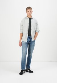 Joven de pie con la mano en el bolsillo, vistiendo una camisa de botones gris claro sobre una camiseta negra, jeans azules y zapatillas negras con rayas blancas.