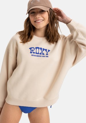 Beige sweatshirt with blue "ROXY" logo, loose fit, ribbed cuffs. Model wearing a light brown cap and blue ribbed swimwear underneath.