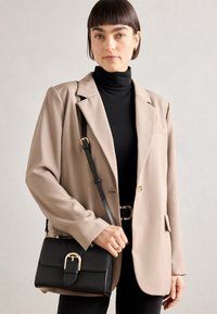 Blazer beige con una vestibilità rilassata, abbinato a un dolcevita nero. Borsa a tracolla nera con dettaglio in metallo sulla fibbia.