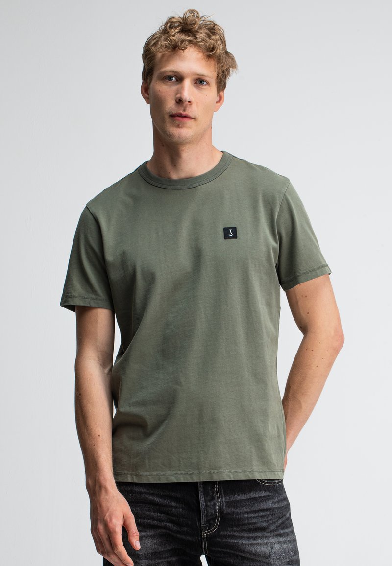 Blonde man in een olijfgroene short-sleeve t-shirt met een klein zwart patch en donkere jeans tegen een plain witte achtergrond.