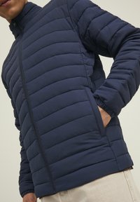Giacca imbottita blu navy con colletto alto, design a righe verticali e chiusura con zip, dotata di tasche laterali e polsini elastici.