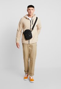 Beige fleece zip-up hoodie med en frontlomme, lysebrune bukser og orange sneakers, bærer en sort crossbody taske med flere rum.