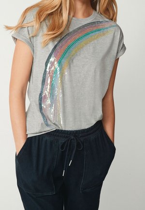 Nainen, joka pukeutunut vaaleanharmaaseen lyhythihaiseen toppiin, jossa värikäs, paljeteilla koristeltu sateenkaarikuvio, sekä tummiin narullisiin housuihin, kädet taskuissa.