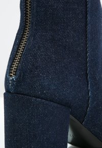 Bottine en denim avec un talon épais, couleur bleu foncé, détail de fermeture éclair sur le côté avec quincaillerie métallique, texture lisse et forme structurée.