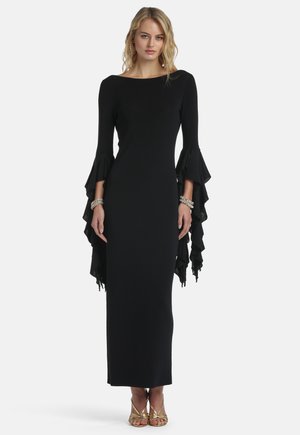 Kobieta w długiej czarnej sukni z rozkloszowanymi, falbaniastymi rękawami, srebrnymi bransoletkami i złotymi sandałami na paski, stojąca na białym tle.