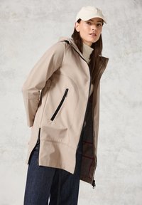 Veste imperméable beige avec capuche, dotée de deux zippers noirs, de poches latérales et d'un ourlet asymétrique. Texture douce et design épuré.