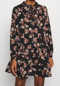 Kwiatowa sukienka z długimi rękawami, kołnierzykiem z wiązaniem oraz warstwowym dołem. Kolory to czerń z różowymi, białymi i pomarańczowymi wzorami kwiatowymi.