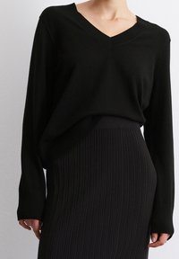 Schwarzer V-Ausschnitt-Pullover mit Rippstruktur, kombiniert mit einem schwarzen Ripp-Rock, der eine lockere Passform und lange Ärmel zeigt.