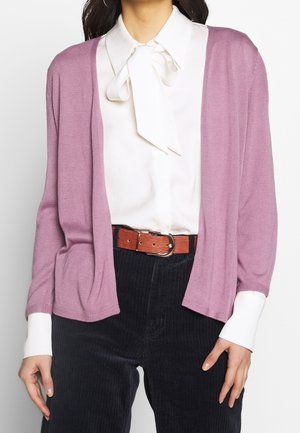 Femme portant un chemisier blanc avec un nœud au col, un cardigan violet clair, une ceinture couleur rouille et un pantalon en velours côtelé noir, debout les mains détendues.