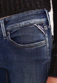 Jeans de mezclilla azul oscuro con un diseño ajustado, que cuentan con un pequeño bolsillo delantero y costuras en contraste. Se puede ver una etiqueta de la marca en la cintura.