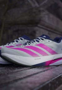 Chaussures de running Adidas blanches et roses avec des accents bleus sur une surface en bois, dotées de la technologie d’amorti LIGHTSTRIKE PRO.