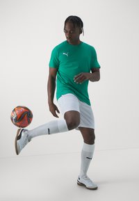 Zelen majica kratkih rukavov za šport z logotipom Puma, bele kratke hlače, bele kolenske nogavice in bela obutev. Nogometna žoga je v gibanju.