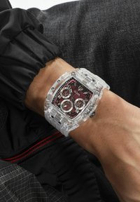 Clear resin watch with a burgundy patterned dial, three subdials, and bold silver accents on a white strap, worn on a wrist.