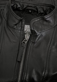 Schwarze Lederjacke mit hohem Kragen, Frontreißverschluss und Steppdetails. Größenschild sichtbar im Nackenbereich. Glatte Textur.