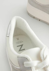 Weiße Sneaker mit einem strukturierten grauen Stoffakzent und metallischen Details. Weiche, plüschige Innenausstattung und gepolsterter Kragen für Komfort. Größenmarkierungen sichtbar.