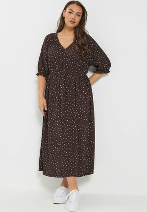 Kvinde stående med hånden på hoften iført en brun midi-kjole med hvide polkaprikker, pufærmer og hvide sneakers foran en ensfarvet baggrund.