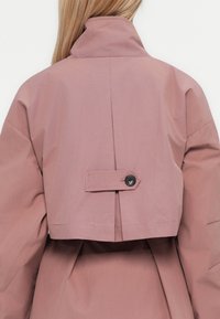 Vue arrière d'une personne portant une veste cropped mauve avec un col haut et un détail de sangle boutonnée au centre du dos.