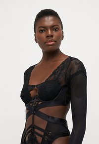 Zwarte lingerie met kantdetails en aansluitende doorzichtige mouwen, met een gestructureerde lijf, satijnen schouderbandaccenten en gouden hardware.