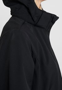 Schwarze Kapuzenjacke mit hohem Kragen, glatter Textur und sichtbaren Nähten. Verfügt über einen durchgehenden Reißverschluss und eine leicht taillierte Passform.