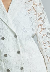 Blazer bianco in pizzo con motivo floreale, caratterizzato da un design doppiopetto e bottoni in tonalità argento, che mette in mostra una texture trasparente e dettagli intricati.