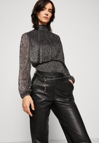 Blouse noire métallisée avec des manches longues en tissu transparent et un col montant, associée à un pantalon en cuir noir taille haute orné d'une chaîne décorative à la taille.