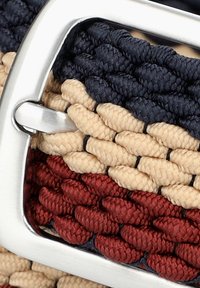 Pleciony pasek z czerwonymi, beżowymi i granatowymi sekcjami oraz błyszczącą srebrną metalową klamrą. Teksturowany wzór z plecionymi motywami.