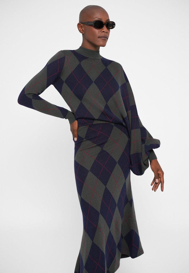 Gebreide jurk in donkergroen en marineblauw met een ruitpatroon en rode accenten. Heeft een hoge hals en lange mouwen met gerimpelde manchetten.